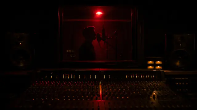 A singer silhouetted at a studio microphone behind vocal-booth glass, lit by a glowing red recording lamp above a mixing console