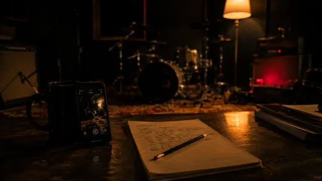 A phone propped against a coffee mug recording a dim rehearsal room, with a handwritten arrangement notebook in the foreground and a drum kit behind